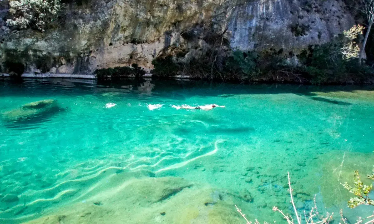 Persona nada en un pou clar de aguas turquesas, bajo un acantilado rocoso.