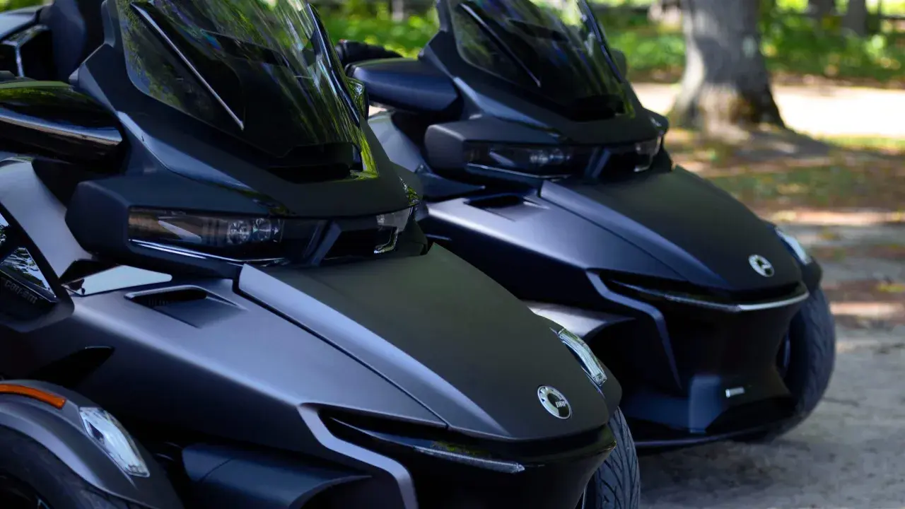 Dwa matowe, czarne pojazdy typu Can-Am Spyder stoją obok siebie na parkingu dla motocykli, gotowe do jazdy.