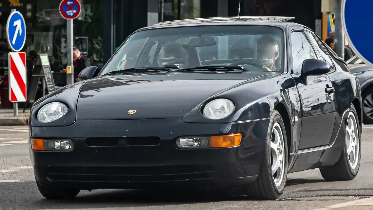 Dunkelblauer 968 Porsche fährt auf der Straße. Ein Mann sitzt am Steuer. Verkehrsschilder sind im Hintergrund zu sehen.