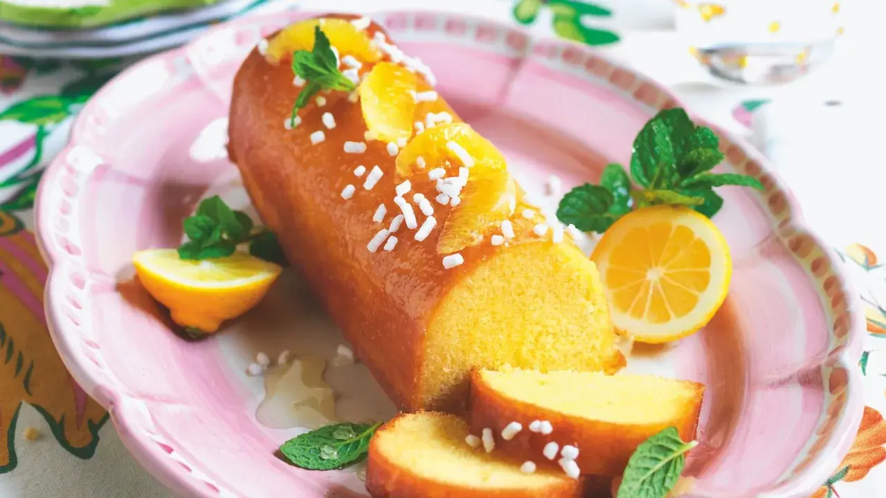 Torta al limone leggerissima, lucida e decorata con fettine d'arancia e zuccherini, servita su un piatto rosa con spicchi di limone e menta.