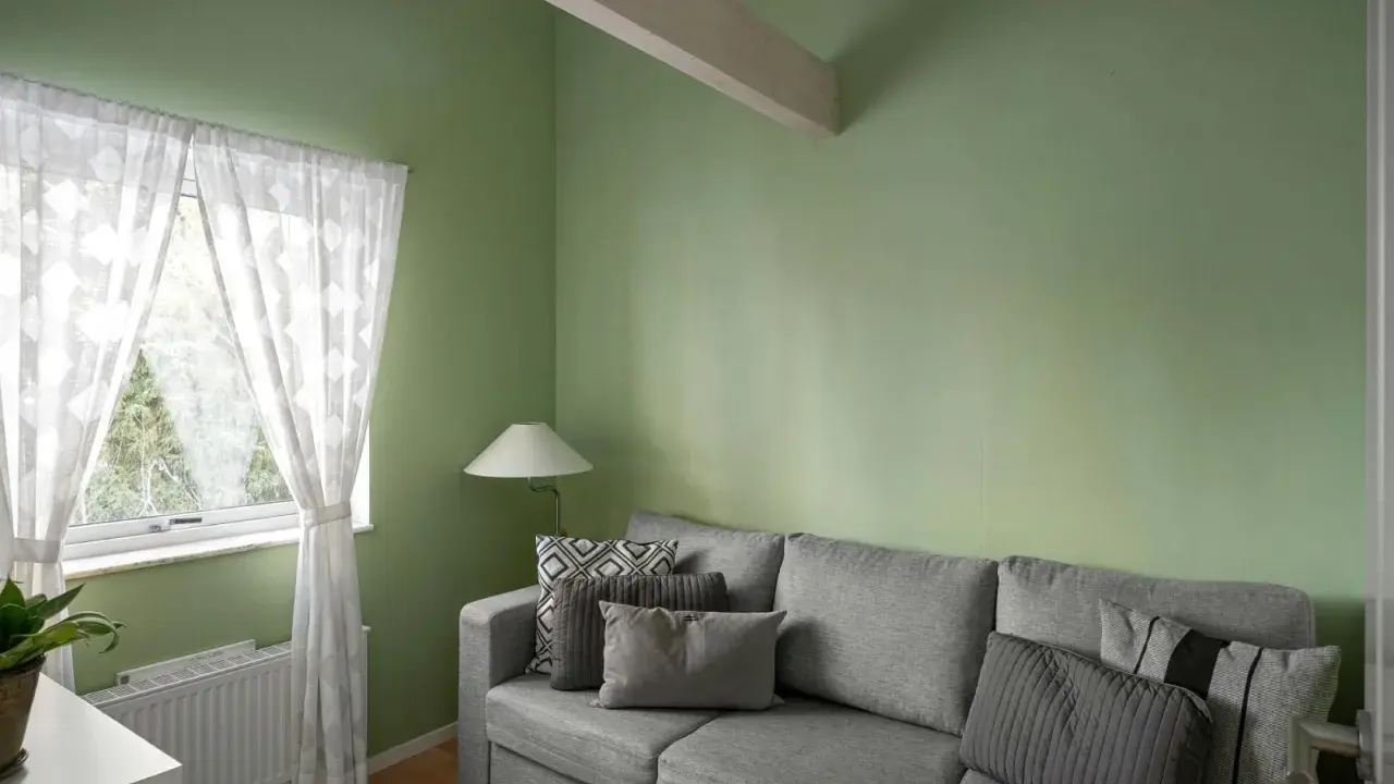 Sofá gris con cojines, lámpara y cortinas blancas junto a pared verde. ¡Excelentes ideas pintar casa con este tono fresco!