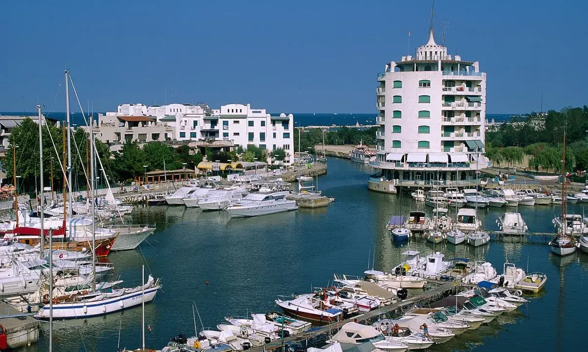 Un vivace porto verde con barche a vela e yacht ormeggiati, edifici bianchi e un edificio circolare a torre.