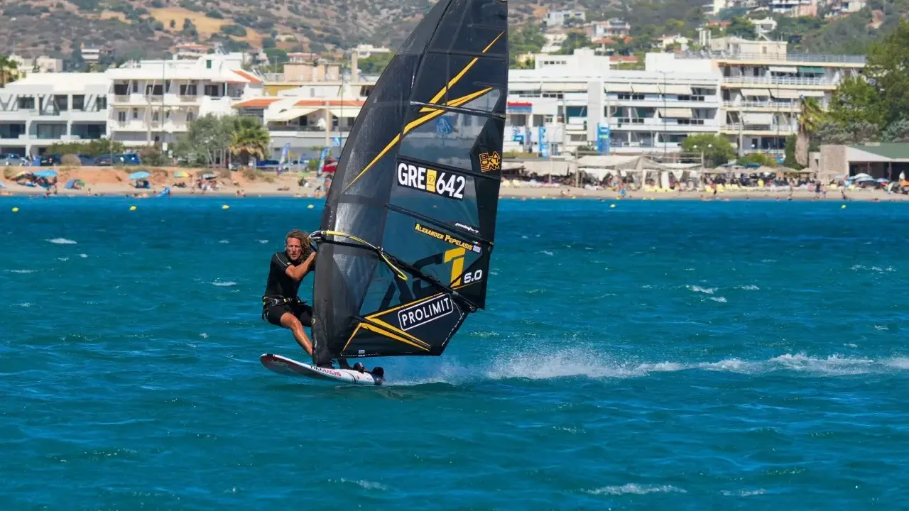 Windsurfer mit schwarzem Segel und gelben Akzenten auf türkisfarbenem Wasser vor einer Küste mit Gebäuden und Bergen. Surfen in Griechenland.