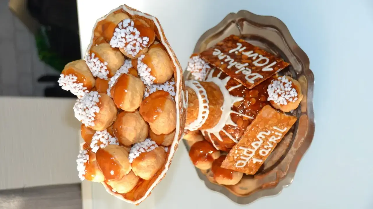 Une pièce montée de choux garnis de crème, nappés de caramel et décorés de perles blanches. Sur un plateau, d'autres choux et des tuiles aux amandes portent des inscriptions.