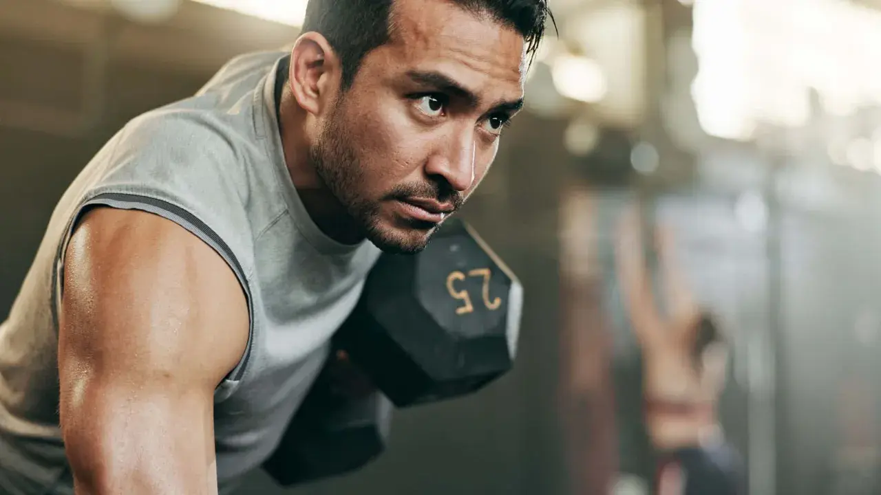 Fokusierter Mann hebt Hantel. Sein Schweiß glänzt. Ein Teil eines 2er Split Trainingsplans.