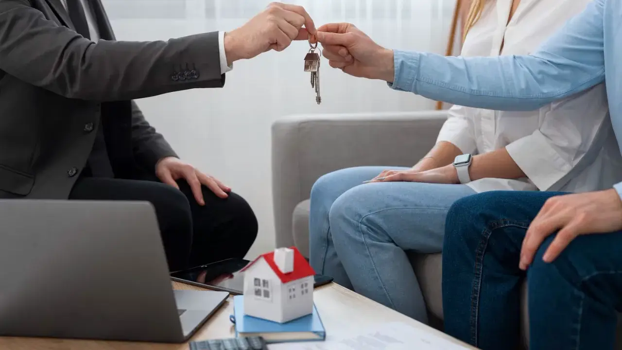 Agente inmobiliario entrega llaves a pareja. Un modelo de casa y calculadora sugieren una transacción, no una notificación de lanzamiento de desahucio.