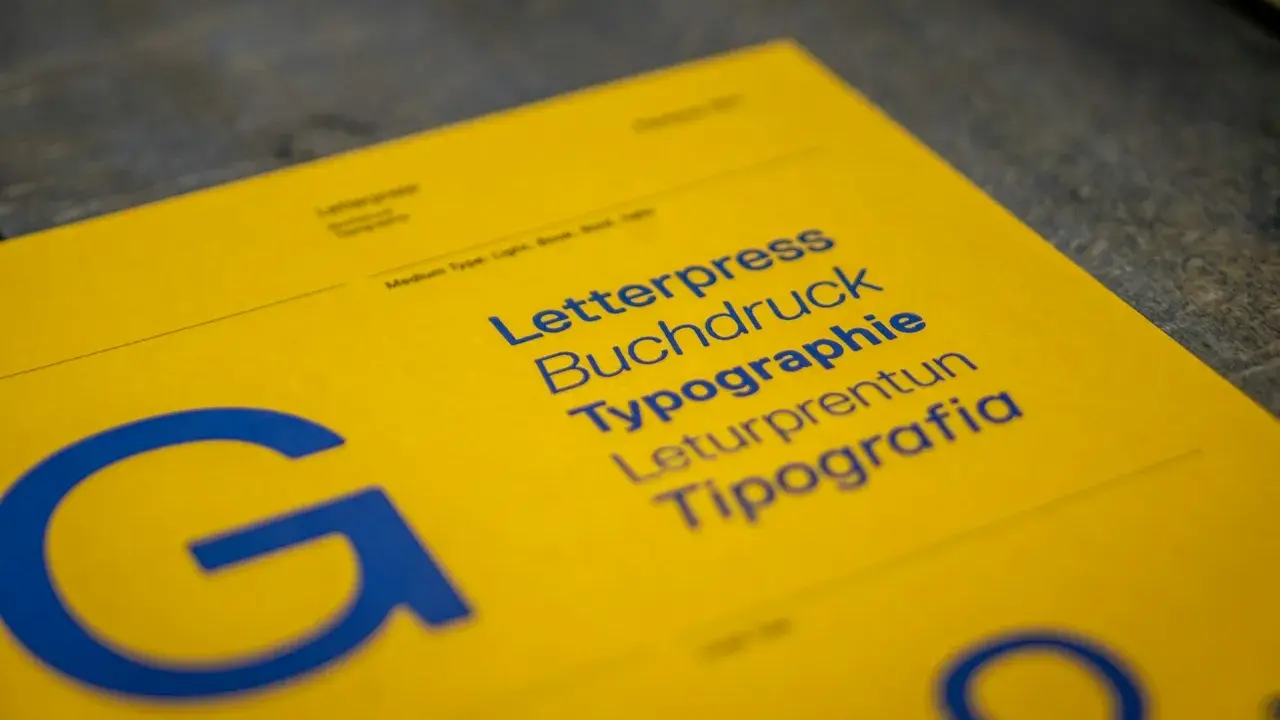 Żółta kartka z niebieskimi napisami: Letterpress, Buchdruck, Typographie, Leturpretun, Tipografia. Duża litera "G" po lewej. To praca typografa.