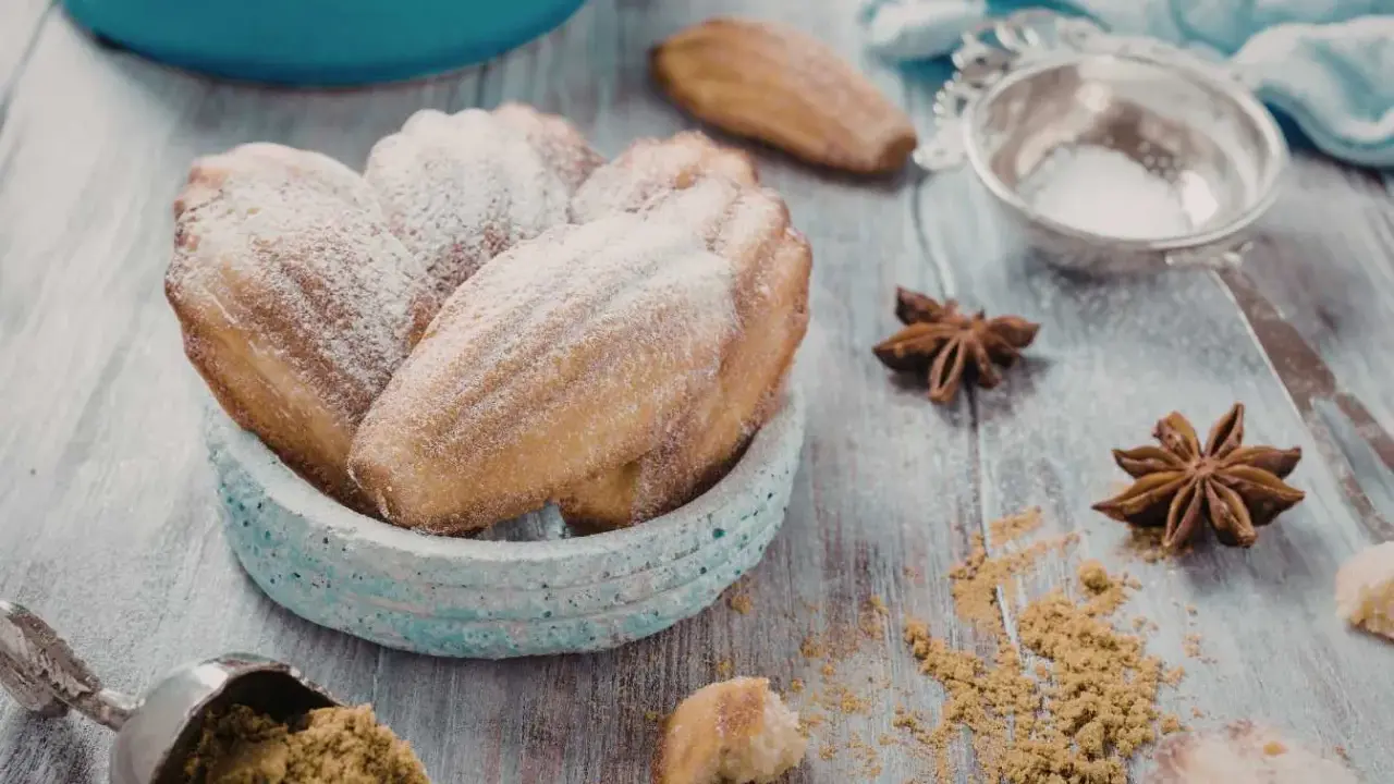 Un bol de biscuits madeleine noisette saupoudrés de sucre glace, avec des étoiles de badiane et une cuillère de poudre sur une table en bois.