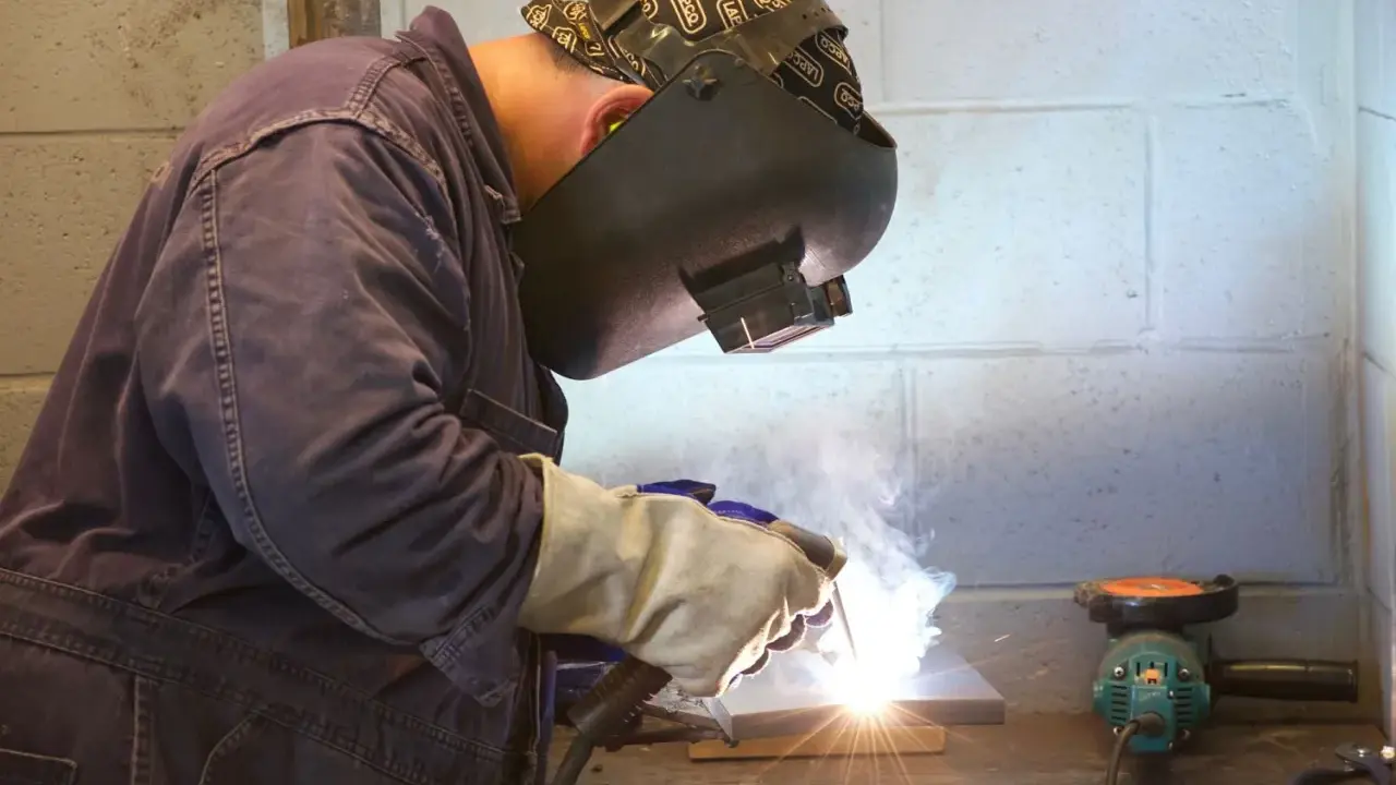 Soldador trabajando con un electrodo, creando chispas y humo. Se aprecian partes del electrodo en acción.