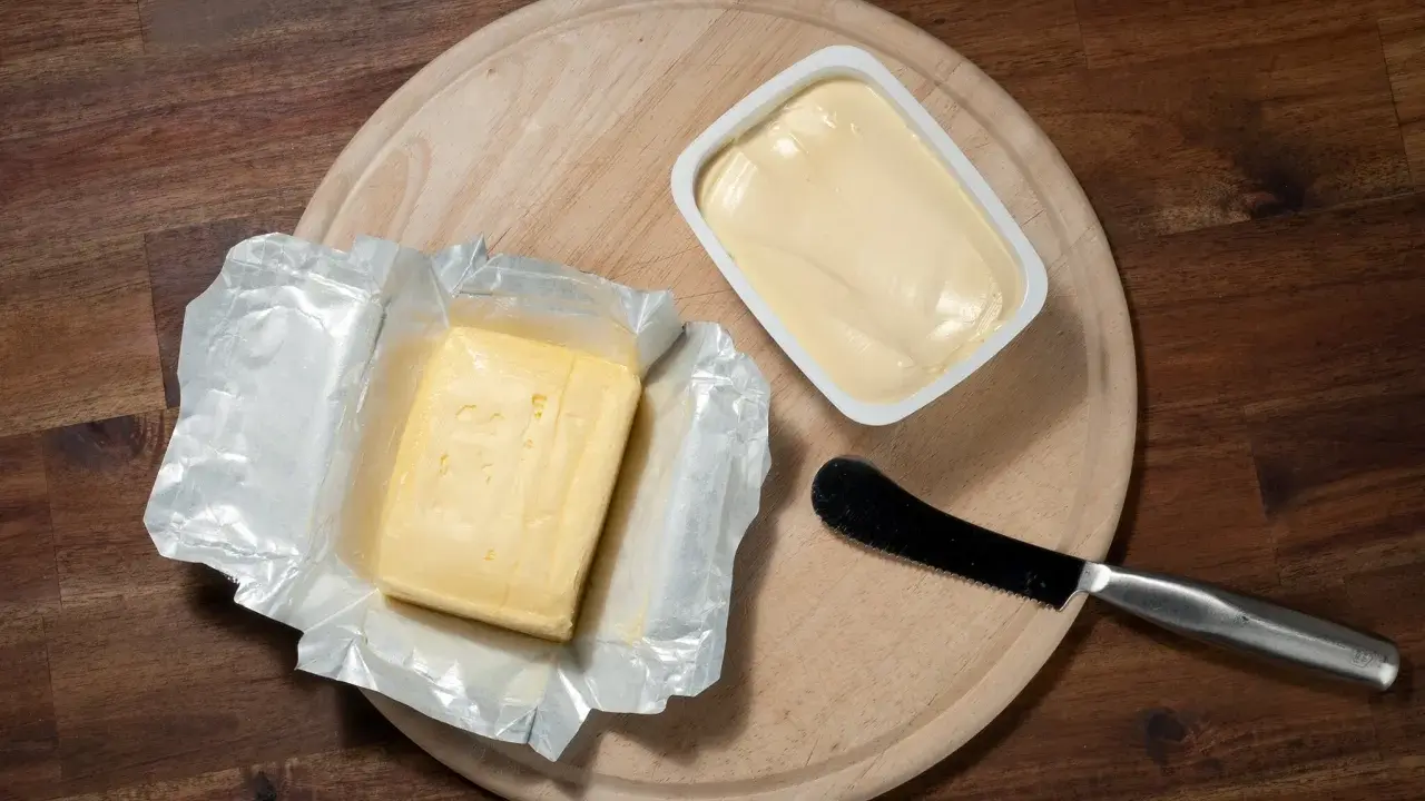Zwei Butterpackungen, eine in Folie, die andere in einer weißen Schale, liegen auf einem Holzbrett. Eine leichte Butter weniger Fett für den gesunden Genuss.