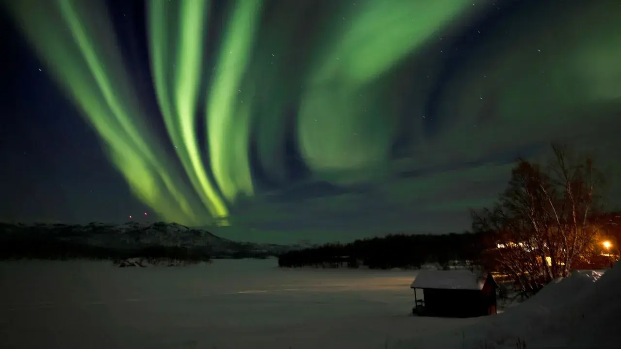 Grön aurora sweden dansar över ett snötäckt landskap med en liten stuga i förgrunden.
