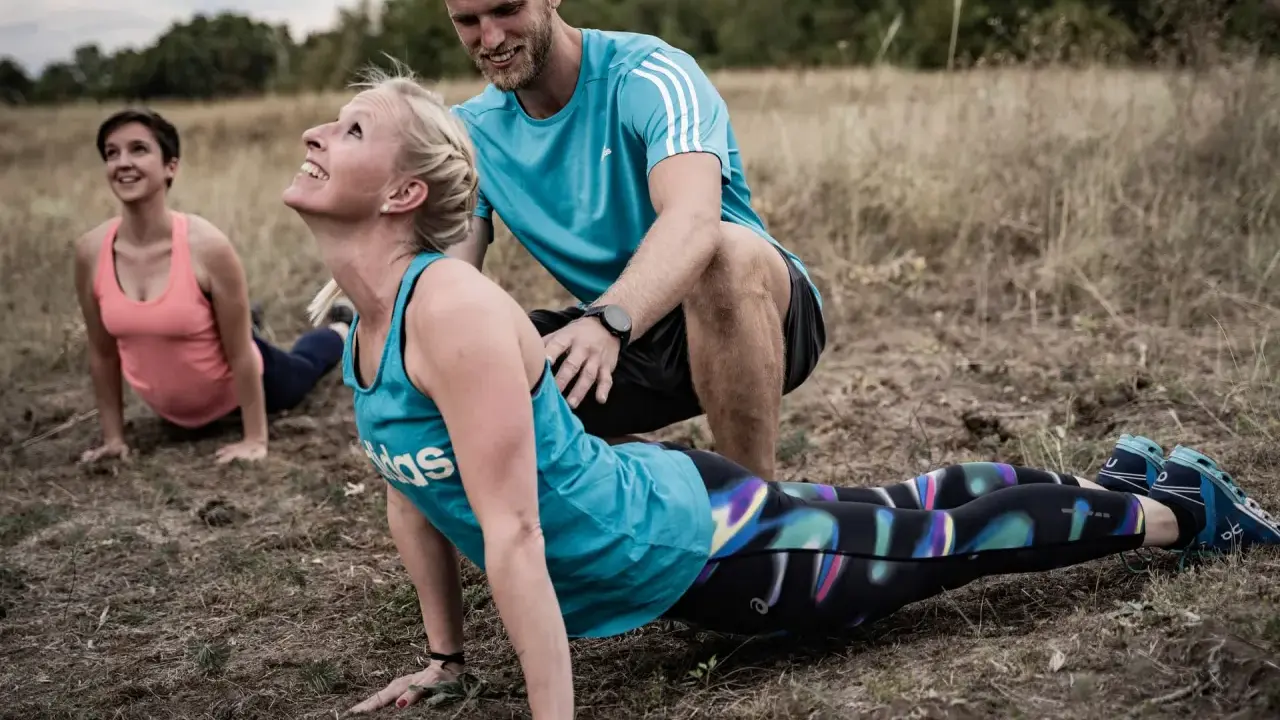 Ein Trainer unterstützt eine Frau bei einer Yoga-Übung im Freien. Das **Faszien Training** stärkt die Körpermitte.