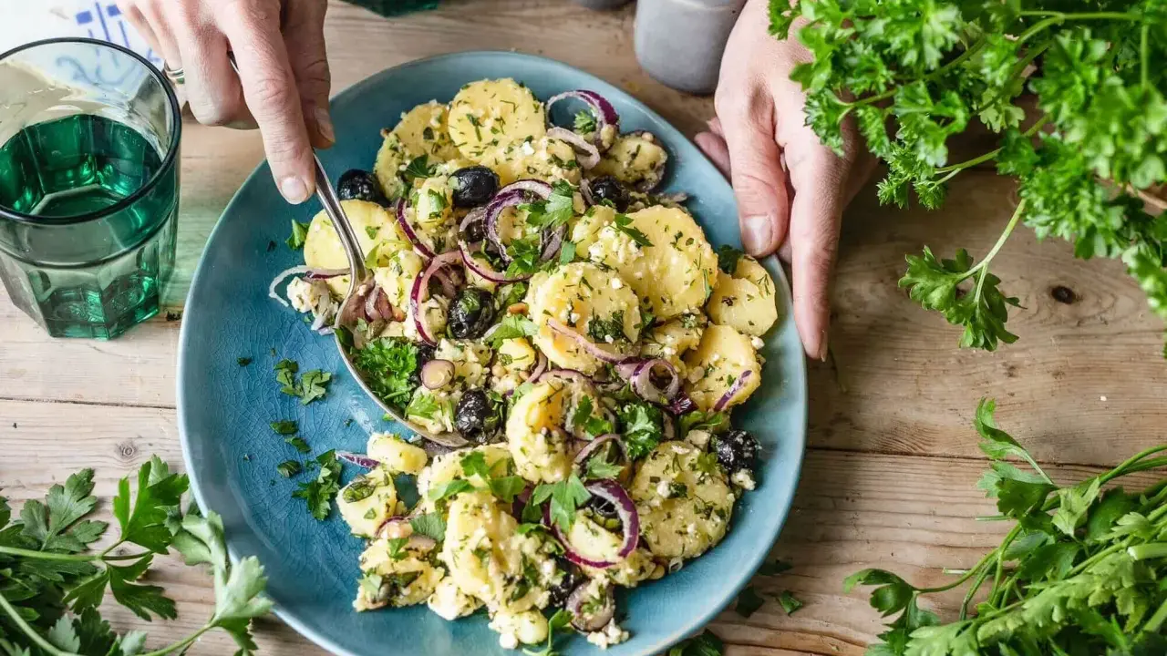 Ein köstlicher mediterraner Kartoffelsalat mit Oliven, roten Zwiebeln und Feta auf einem blauen Teller, garniert mit frischer Petersilie.
