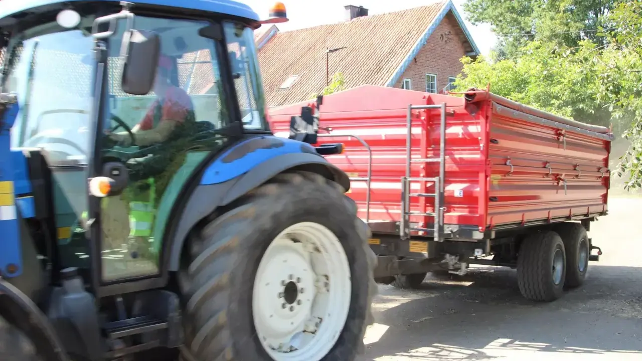 Rolnik z prawem jazdy na ciągnik prowadzi maszynę z czerwoną przyczepą.