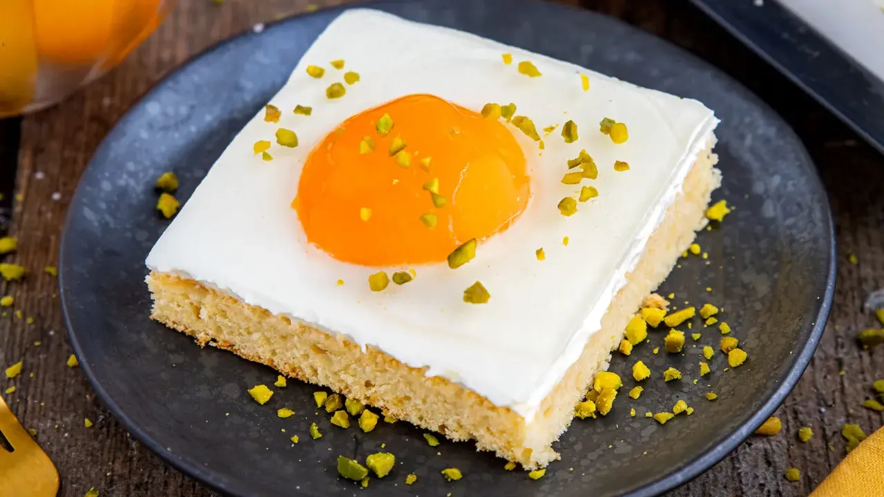 Ein Stück Kuchen mit weißer Glasur, einem orangen Gelee-Dotter und gehackten Pistazien, das wie ein Spiegeleikuchen mit Quark aussieht.