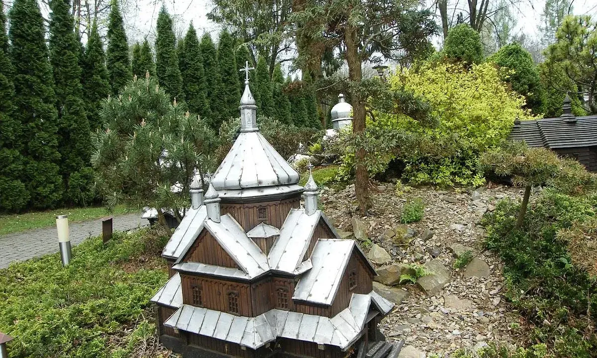 Drewniana cerkiewka z metalowym dachem, miniatura w parku miniatur Myczkowce, otoczona zielenią i kamieniami.