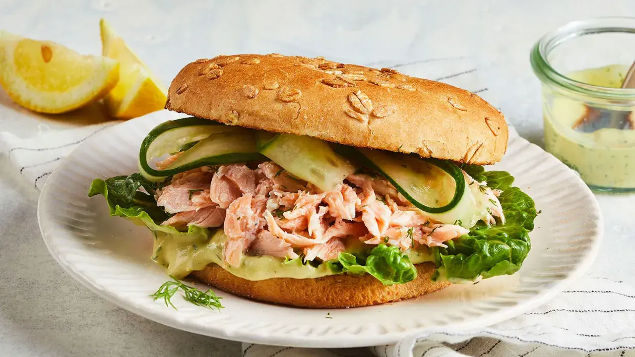Ein Brötchen mit pulled Lachs, Salat, Gurkenscheiben und einer cremigen Sauce. Dazu Zitronenspalten und ein Dip.
