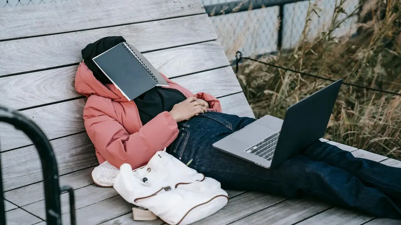 Osoba w różowej kurtce leży na ławce, z zeszytem na twarzy. Obok leży laptop i plecak. Czas na odpoczynek i dodanie nowych pomysłów.