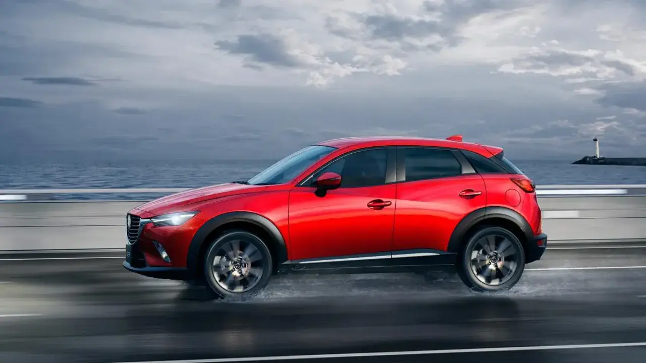 Un Mazda CX-3 rojo circula por una carretera mojada junto al mar. Las medidas del Mazda CX-3 son ideales para la ciudad y la aventura.