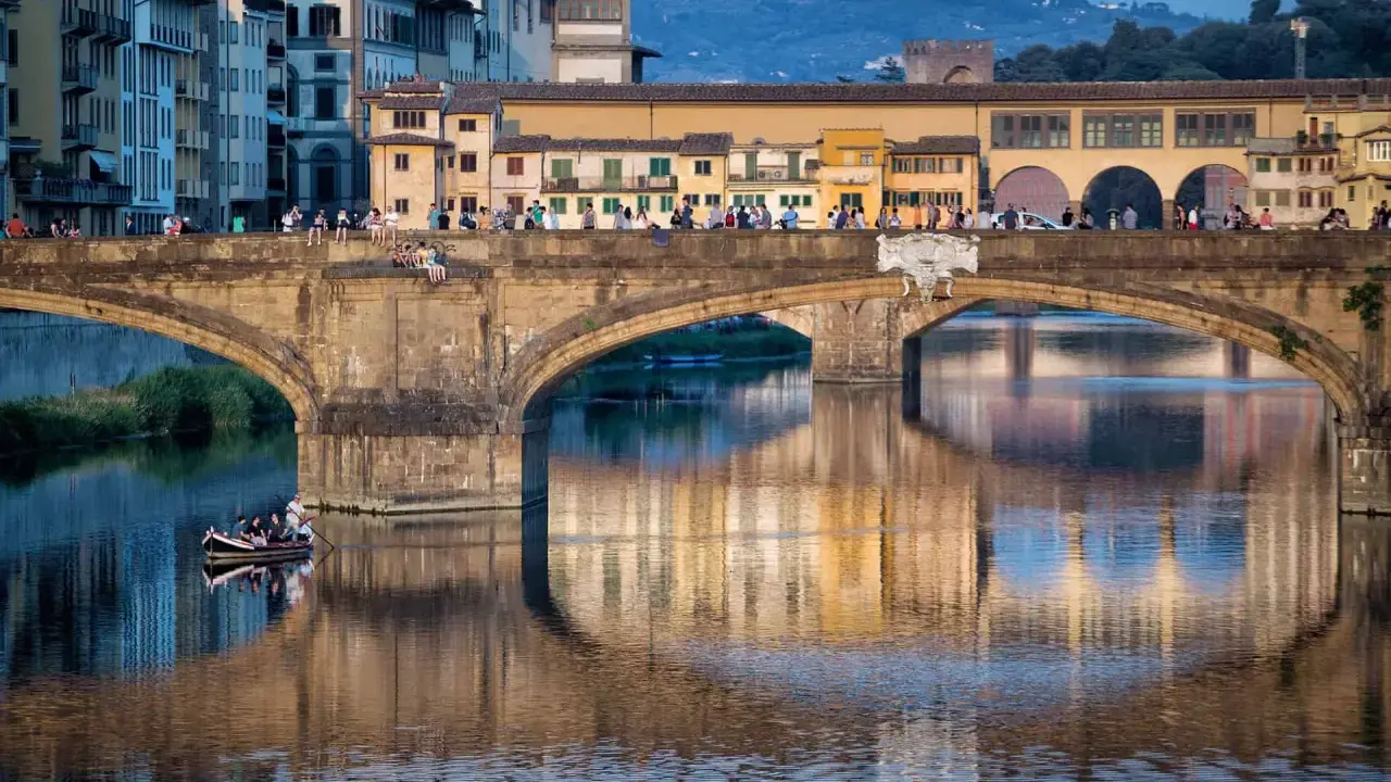 Le Ponte Vecchio à Florence, avec ses maisons colorées et ses reflets sur l'Arno. Une barque navigue paisiblement sous le pont.