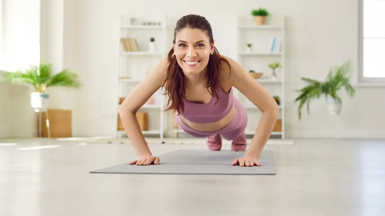 Frau in lila Sportkleidung macht Liegestütze für Yoga Muskelaufbau. Sie lächelt glücklich in die Kamera.