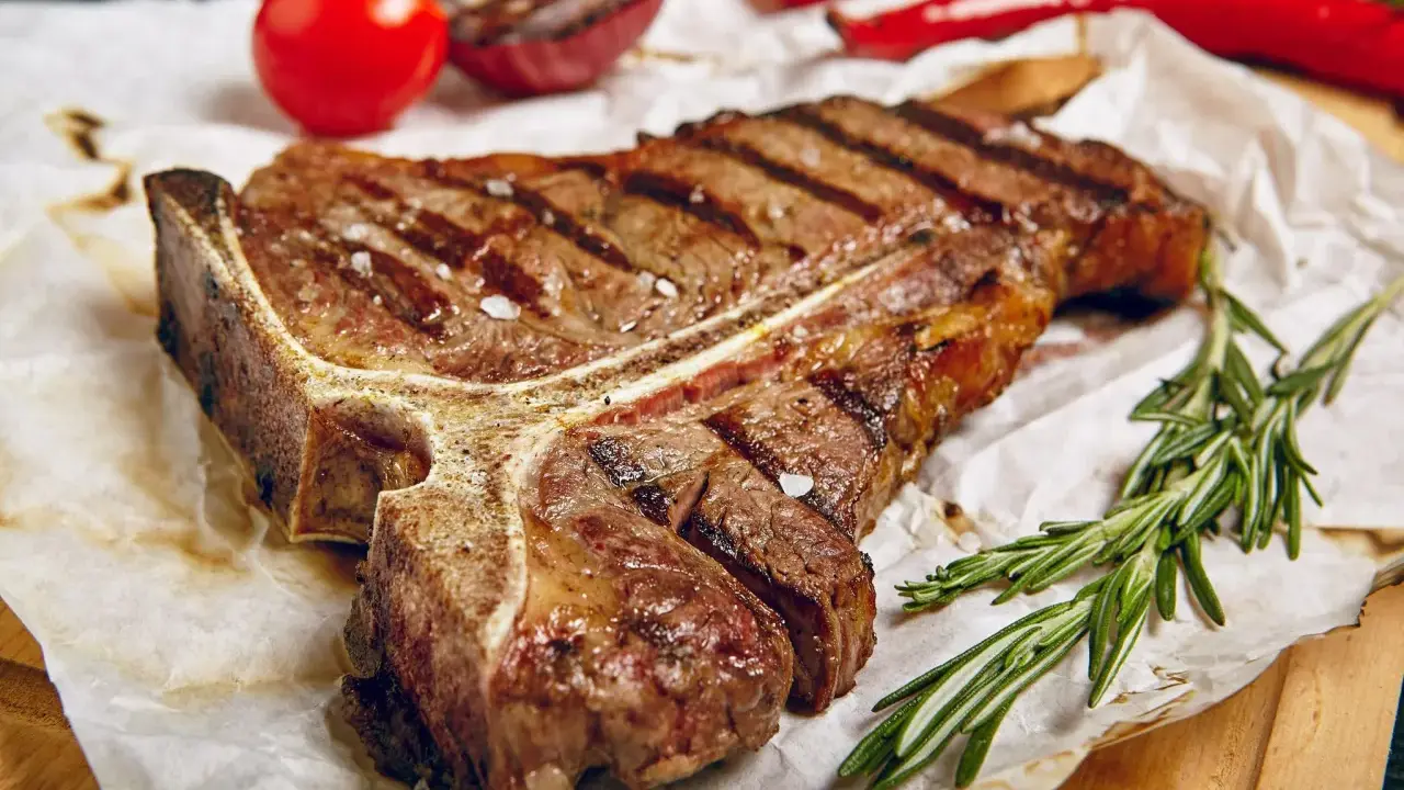 Saftiges T-Bone-Steak, perfekt gegrillt, mit grobem Salz bestreut, daneben Rosmarin, Tomaten und Chili.