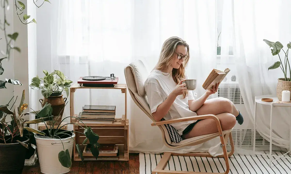Mujer leyendo en un sillón, taza en mano, rodeada de plantas y libros. Un rincón de lectura con decoración acogedora y un tocadiscos.