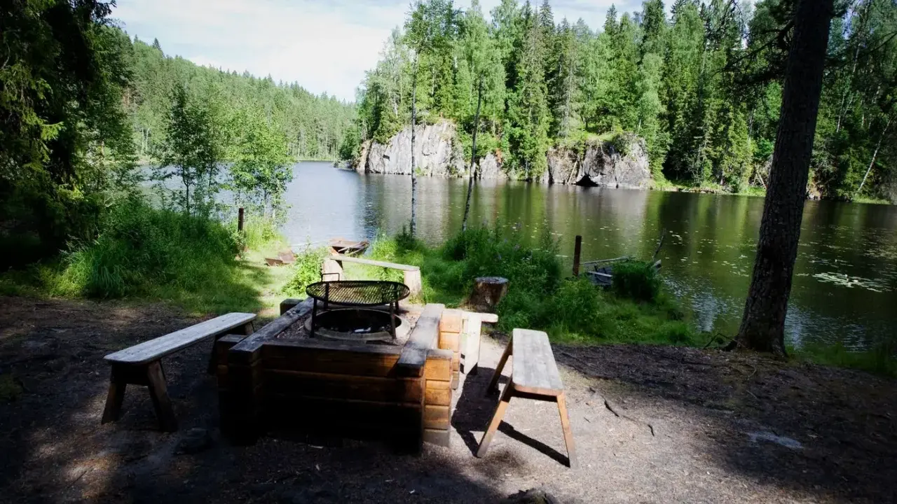 En mysig eldstad med bänkar vid en stilla sjö, omgiven av grönskande skog. Ett perfekt utflyktsmål i Dalarna för avkoppling.