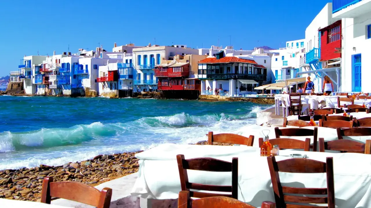 Cosa vedere a Mykonos in crociera: case colorate sul mare, onde che si infrangono sulla spiaggia di ciottoli e tavoli apparecchiati per godersi il panorama.