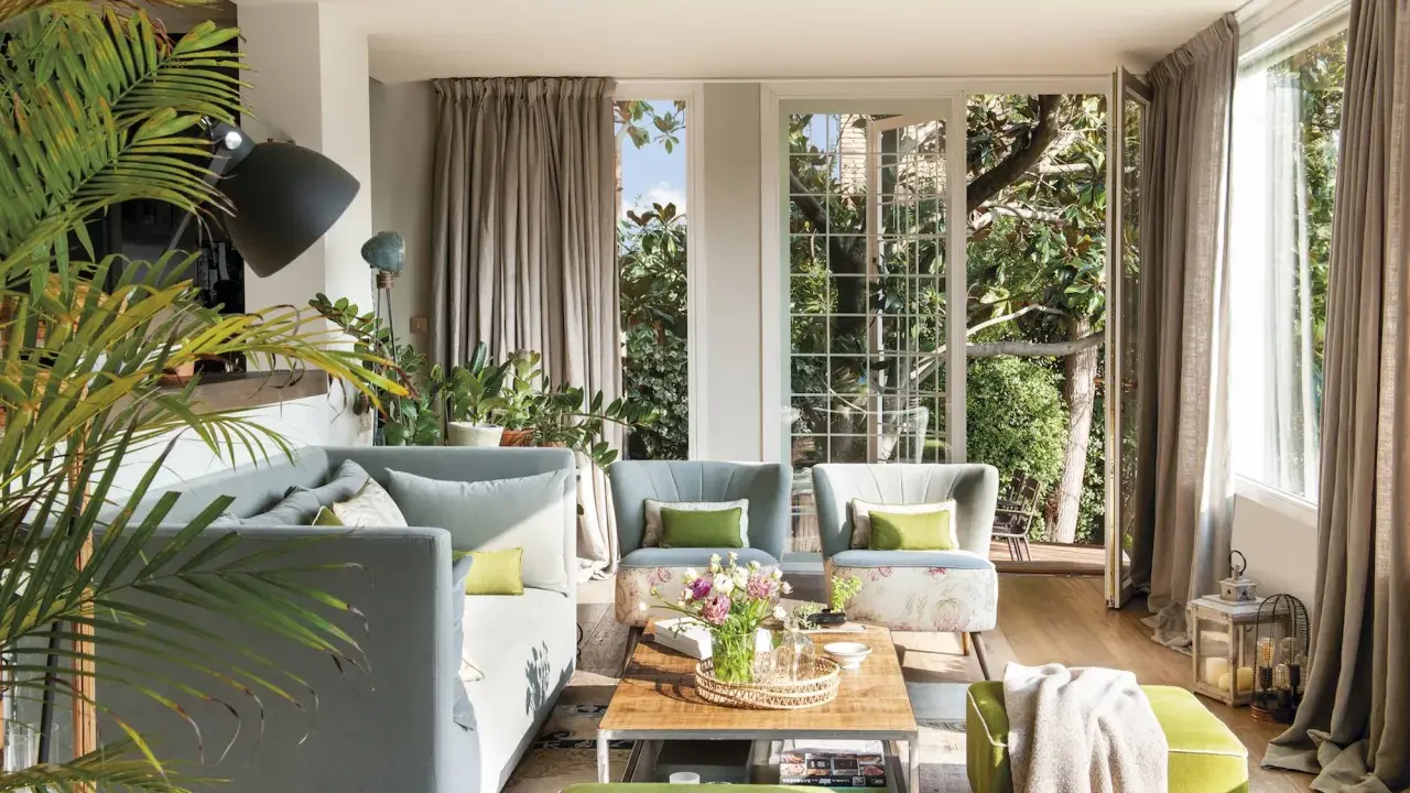Sofá gris y otomanas verdes crean un ambiente acogedor. Plantas exuberantes y luz natural inundan el salón.