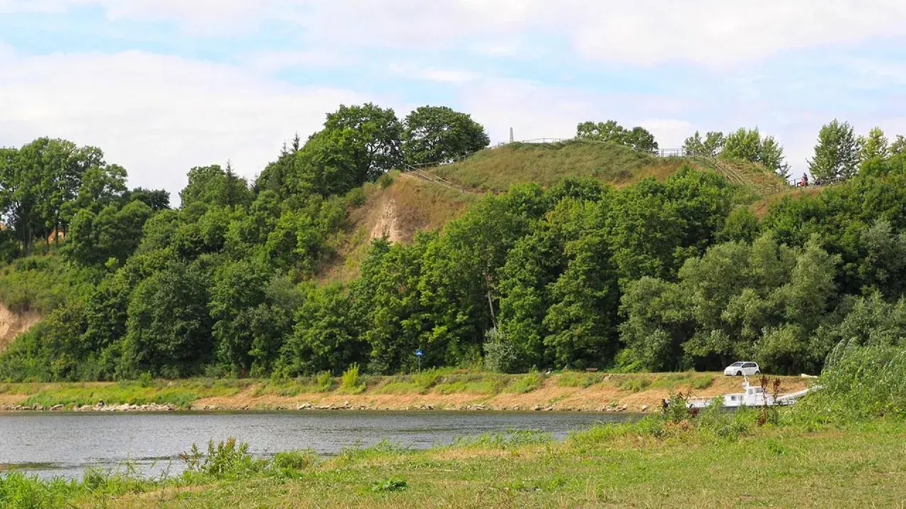 Zielona skarpa nad rzeką, na szczycie której widać ścieżki i ogrodzenie. To góra zamkowa w Drohiczynie, miejsce pełne historii.