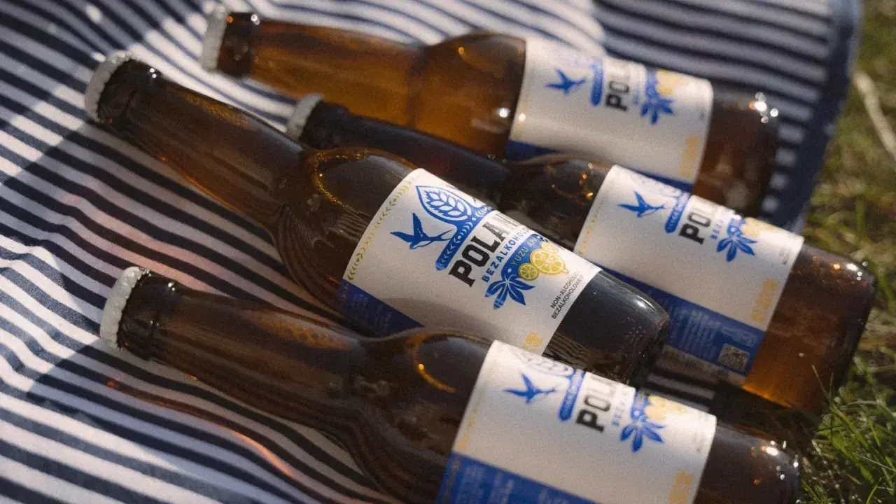 Butelki piwa POLAN z napisem "BEZALKOHOLOWE" leżą na kocu w paski. To piwo ma 0 alkoholu, idealne na upalne dni.