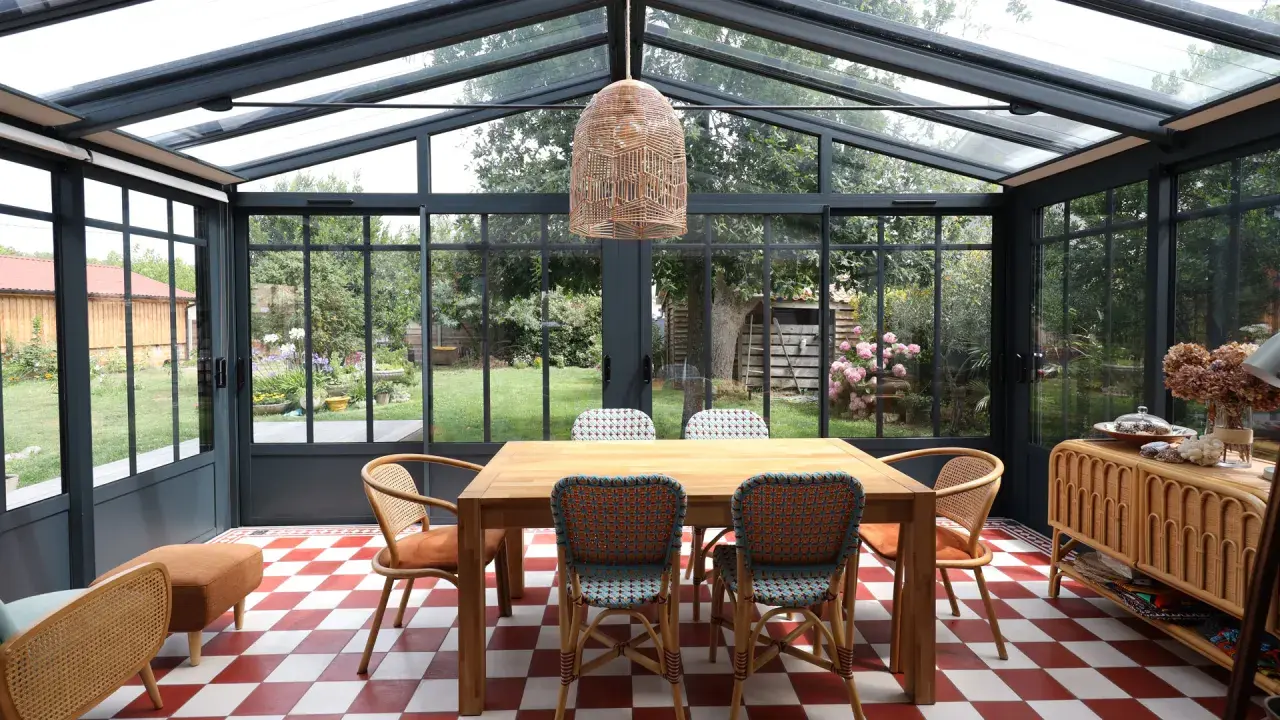 Salle à manger lumineuse dans une véranda avec toiture zinc. Table en bois, chaises colorées, sol à damier rouge et blanc, vue sur jardin verdoyant.