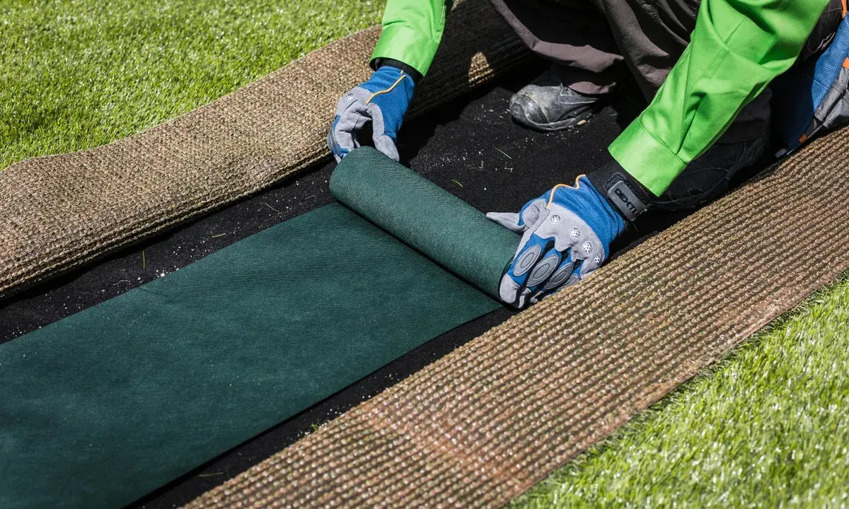 Manos enguantadas desenrollan césped artificial verde sobre una base negra. Paso clave para saber como colocar cesped artificial.