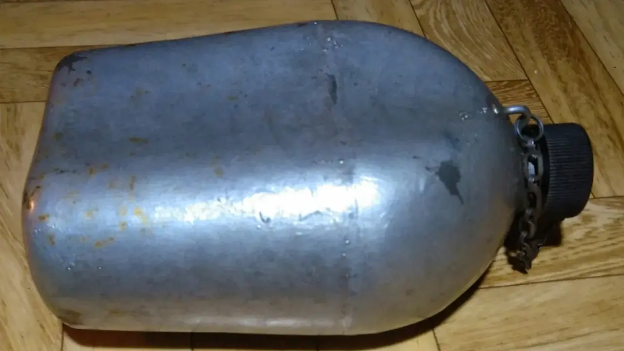 Metalowa manierka US Army z czarną zakrętką i łańcuszkiem. Widoczne ślady użytkowania i rdzy.
