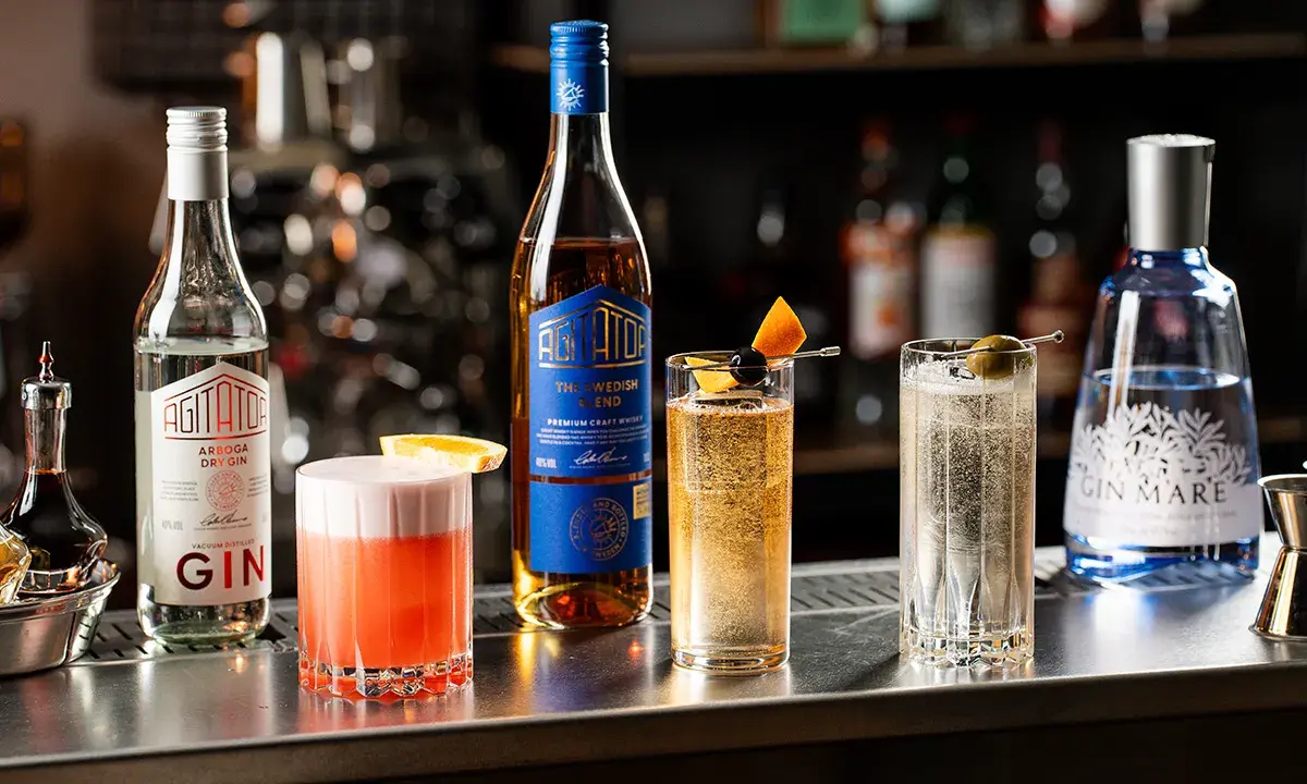 En bar med flera drinkar, inklusive en rosa drink, en gyllene drink med apelsin och körsbär, och en klar drink med oliv. Flera flaskor syns, bland annat en gin och en whisky. En perfekt kväll med en god bacardi drink.