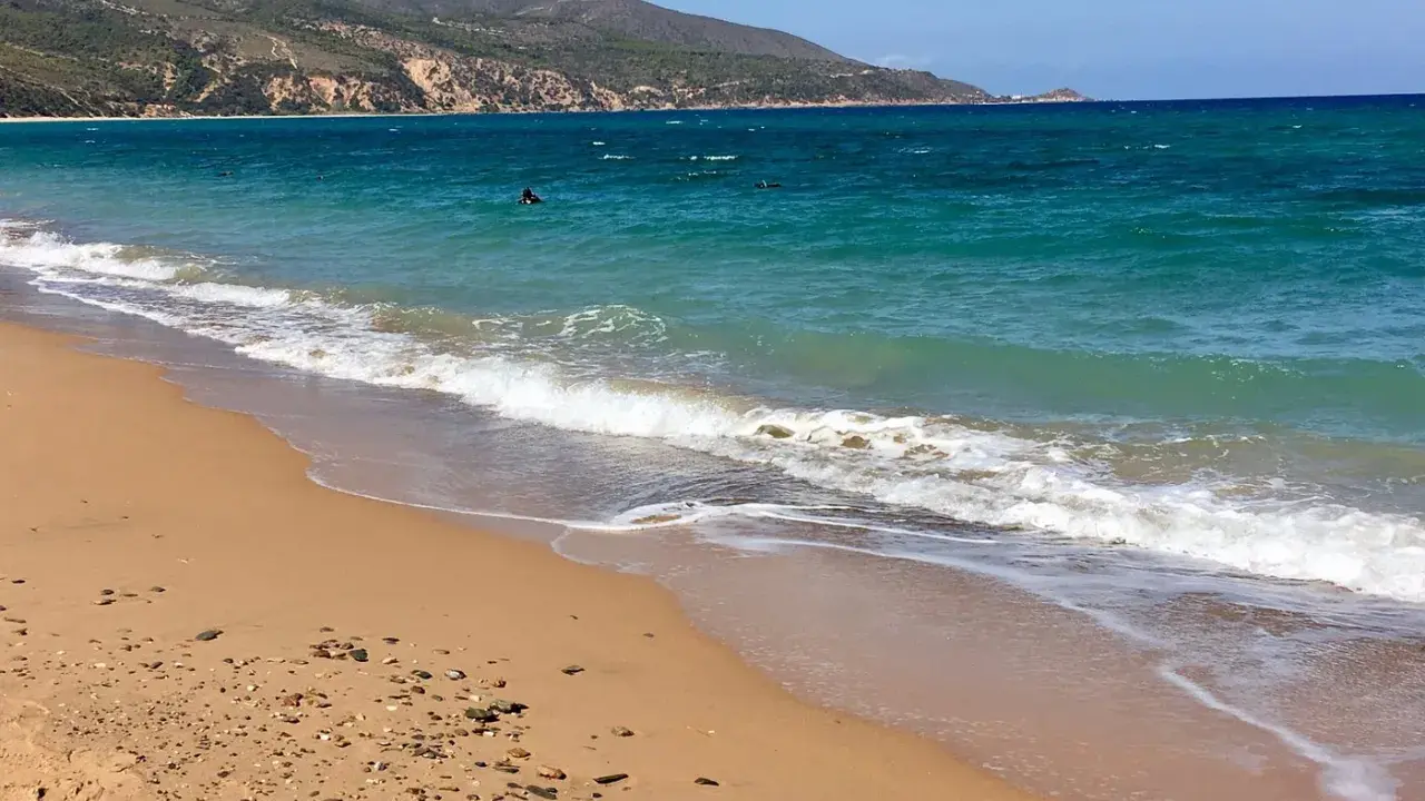 Türkisblaues Meer und ein sandiger Strand bei Oran Stadt. Wellen brechen sanft, im Wasser sind einige Schwimmer zu sehen.
