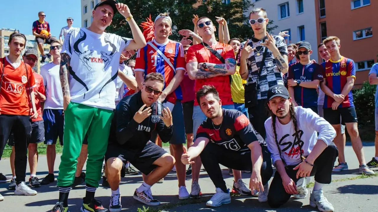 Grupa młodych ludzi w strojach piłkarskich, pozująca do zdjęcia. W tle budynki mieszkalne. To świetny **reto tekst** dla fanów futbolu.