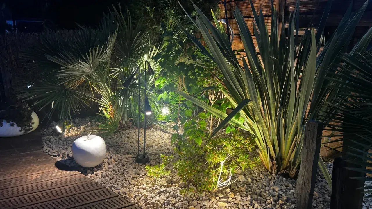 Jardin illuminé la nuit, créant une ambiance chaleureuse. La **norme éclairage extérieur** est respectée pour mettre en valeur la végétation luxuriante et le chemin en bois.
