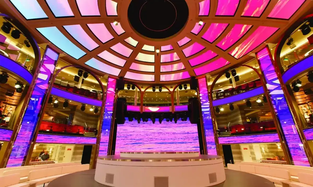 Interni di una nave da crociera, con un palco illuminato da luci rosa e blu, che ricorda la costa mediterranea.
