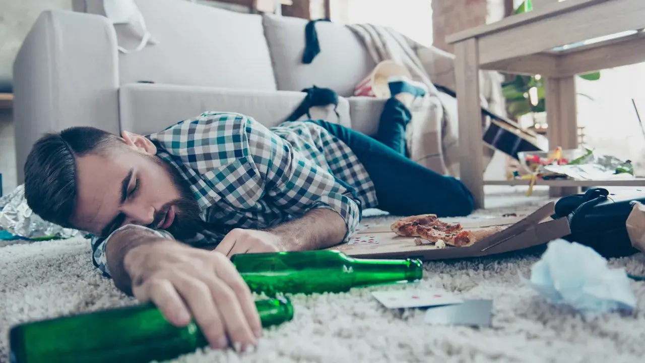 Mężczyzna śpi na dywanie, otoczony pustymi butelkami i resztkami jedzenia. To obrazek pokazujący skutki nadużywania alkoholu, jak padaczka alkoholowa po jakim czasie picia może się pojawić.