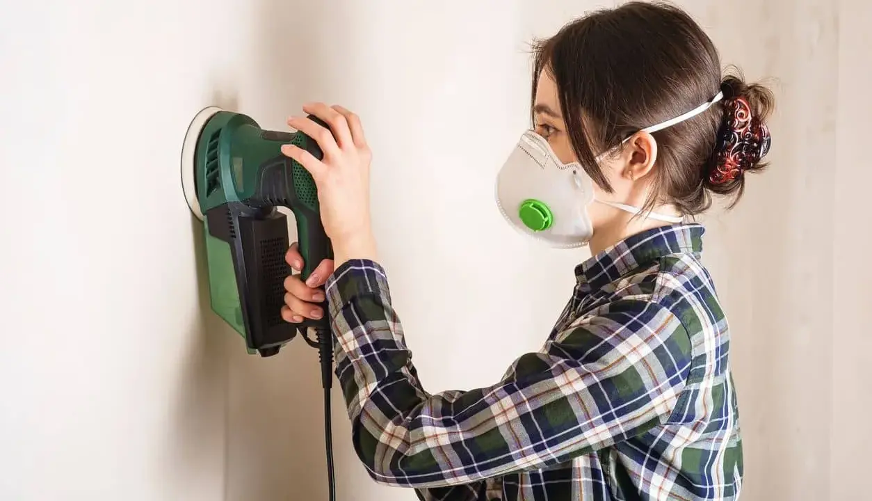 Mujer con mascarilla lijando una pared con una lijadora eléctrica.