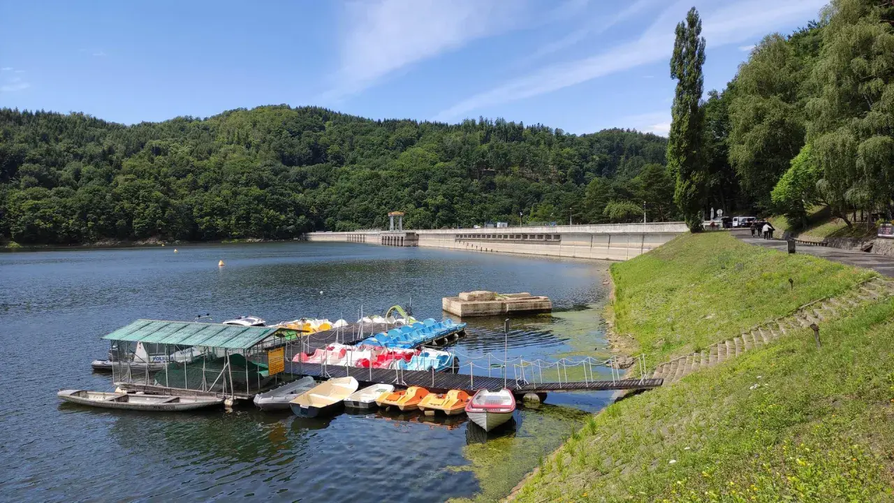 Jezioro Rożnowskie zaprasza na wodną trasę. Na pomoście czekają kolorowe rowerki wodne i łodzie, a w tle wznosi się imponująca zapora.