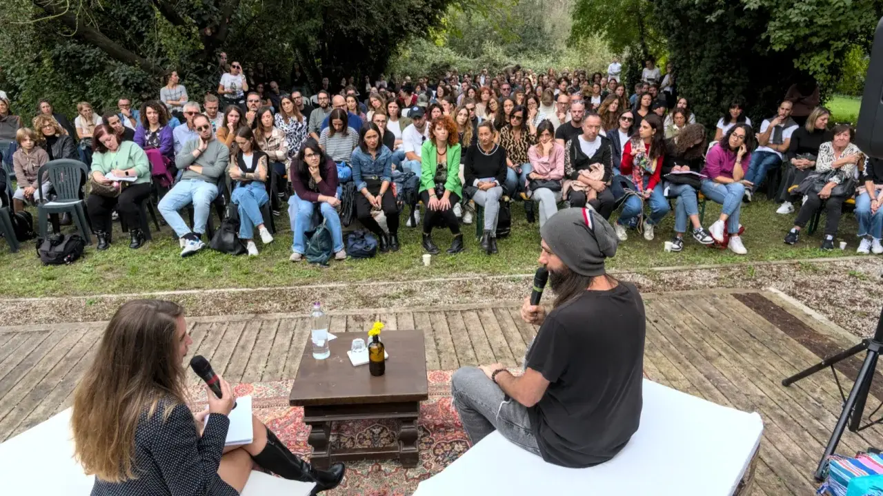 Un pubblico attento ascolta un dibattito all'aperto. Le parole fluiscono, profonde come il mare, leggere come il cielo.