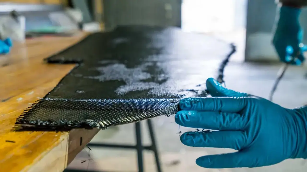 Main d'une personne en gant bleu manipulant un tissu de carbone imbibé de résine, une étape clé pour comprendre qu'est-ce que l'epoxy dans la fabrication composite.