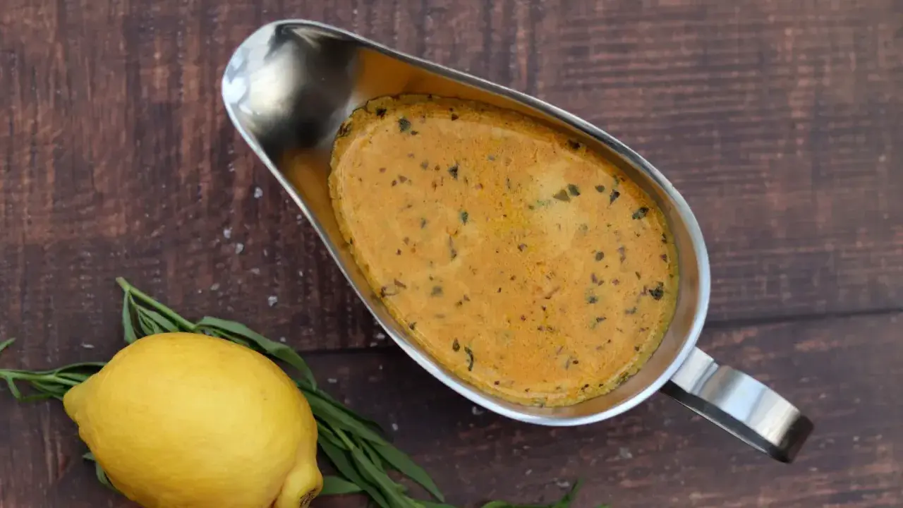 Eine cremige Cafe de Paris Sauce in einer Sauciere, daneben ein Zitrone und Kräuter auf dunklem Holz.