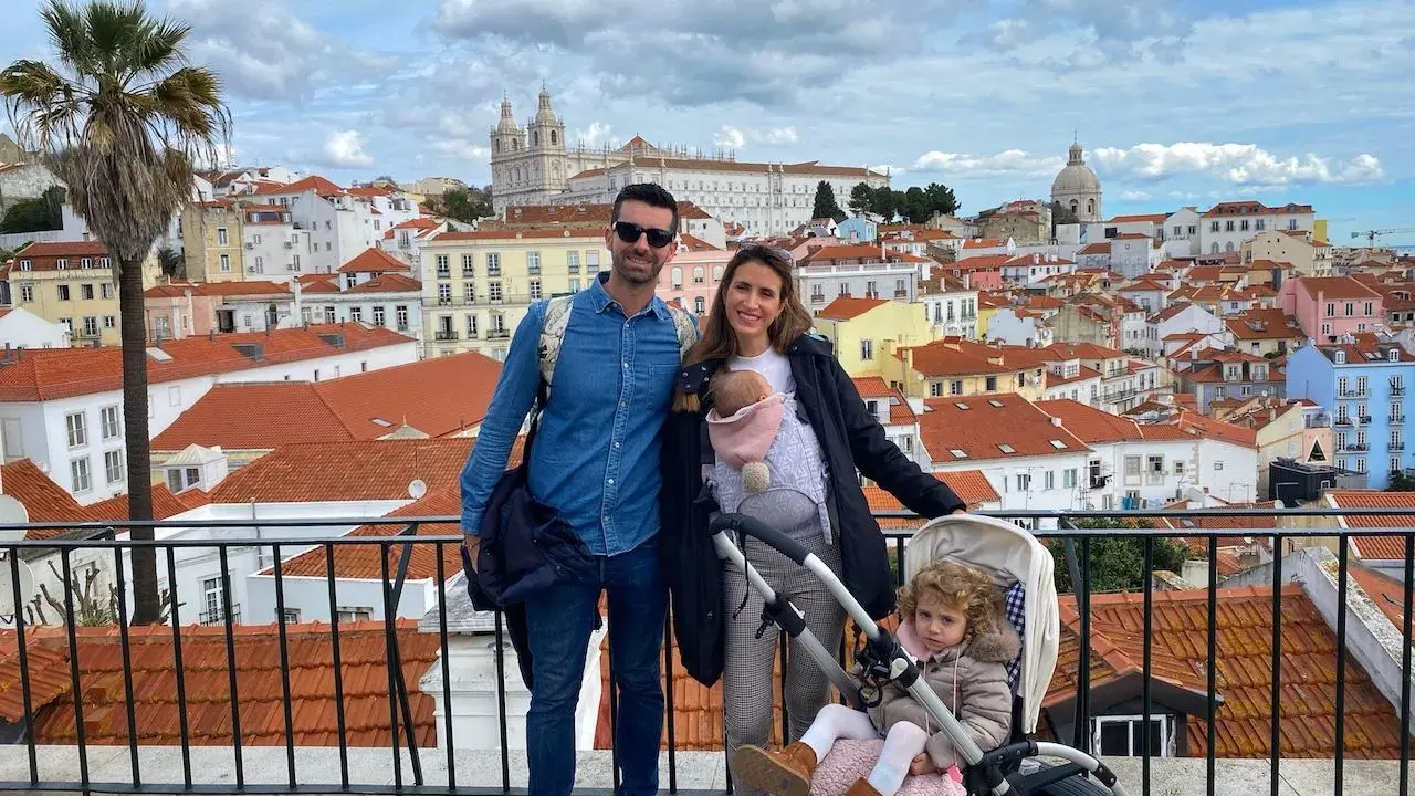 Familia disfrutando de las vistas de Lisboa con niños. Un bebé en portabebés y una niña en carrito, con la ciudad de tejados naranjas de fondo.