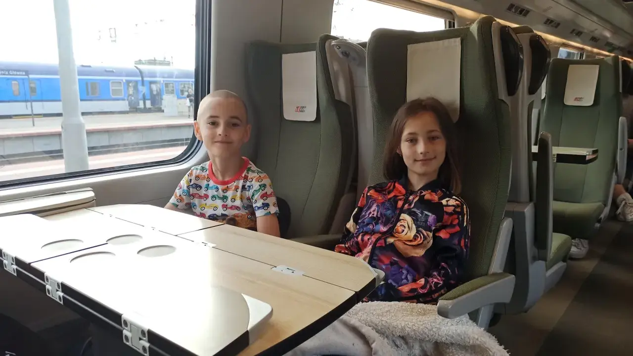 Dzieci siedzą w przedziale dla osoby z dzieckiem do lat 6 Intercity. Chłopiec w kolorowym T-shircie i dziewczynka w kurtce z kwiatowym wzorem.