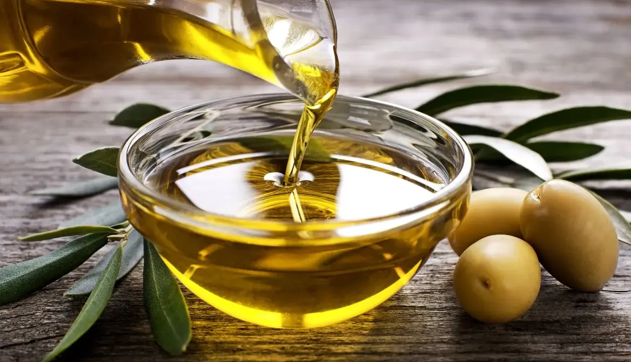 Aceite de oliva virgen extra vertiéndose en un cuenco de cristal, rodeado de aceitunas y hojas de olivo sobre madera rústica.