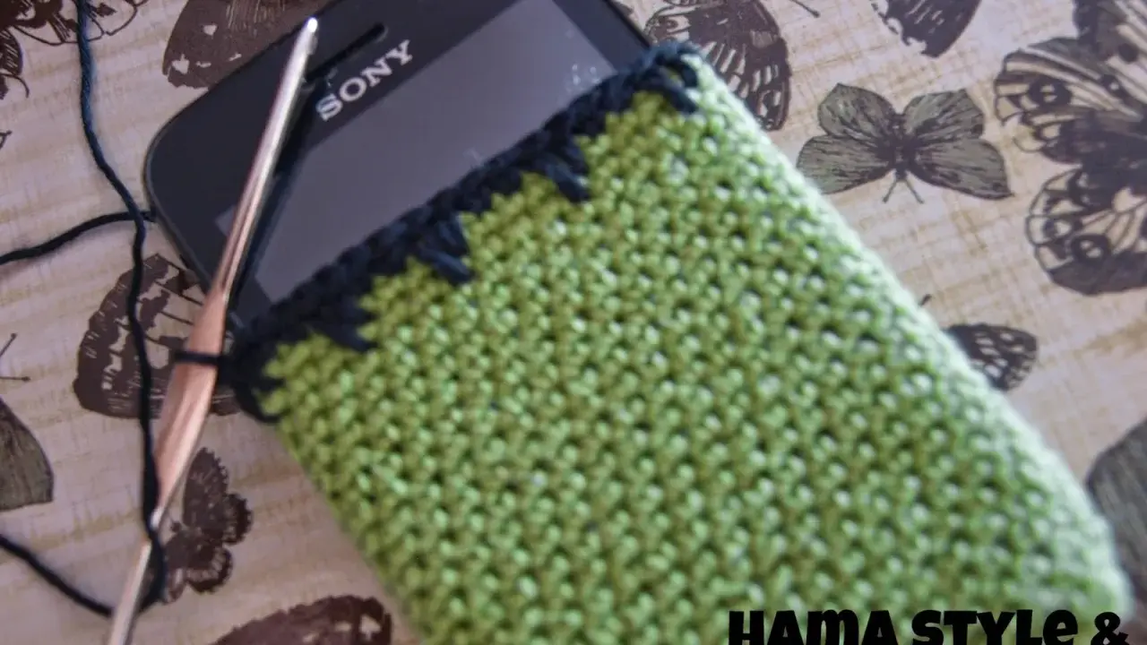 Funda de móvil tejida a crochet, un patrón verde con borde negro. Un gancho y hilo negro están listos para continuar el proyecto.